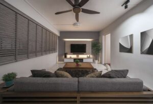 Modern living room with a wall-mounted flat-screen TV, a grey sofa facing it, and a stylish wooden ceiling fan with brown blades. The room blends technology and comfort, creating a contemporary home entertainment space.