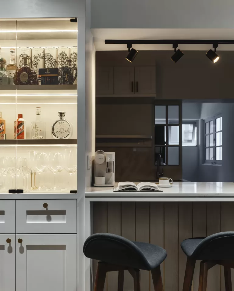 Custom built-in bar cabinet with internal lighting and glass doors next to a white open-concept kitchen counter with warm wood paneling and black stools.