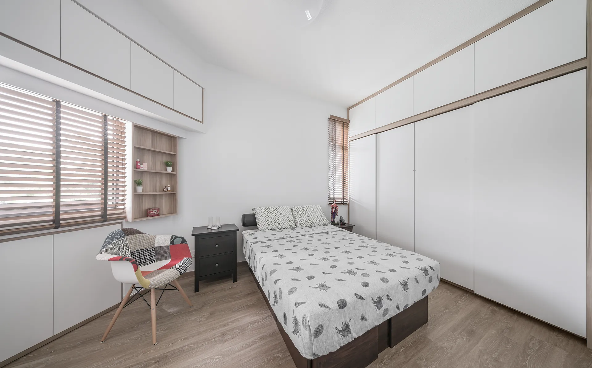 A clean, minimalist bedroom with a low-profile bed and a neutral-toned bedspread. A colorful patchwork accent chair sits near a window with wooden blinds, adding a pop of color to the space.