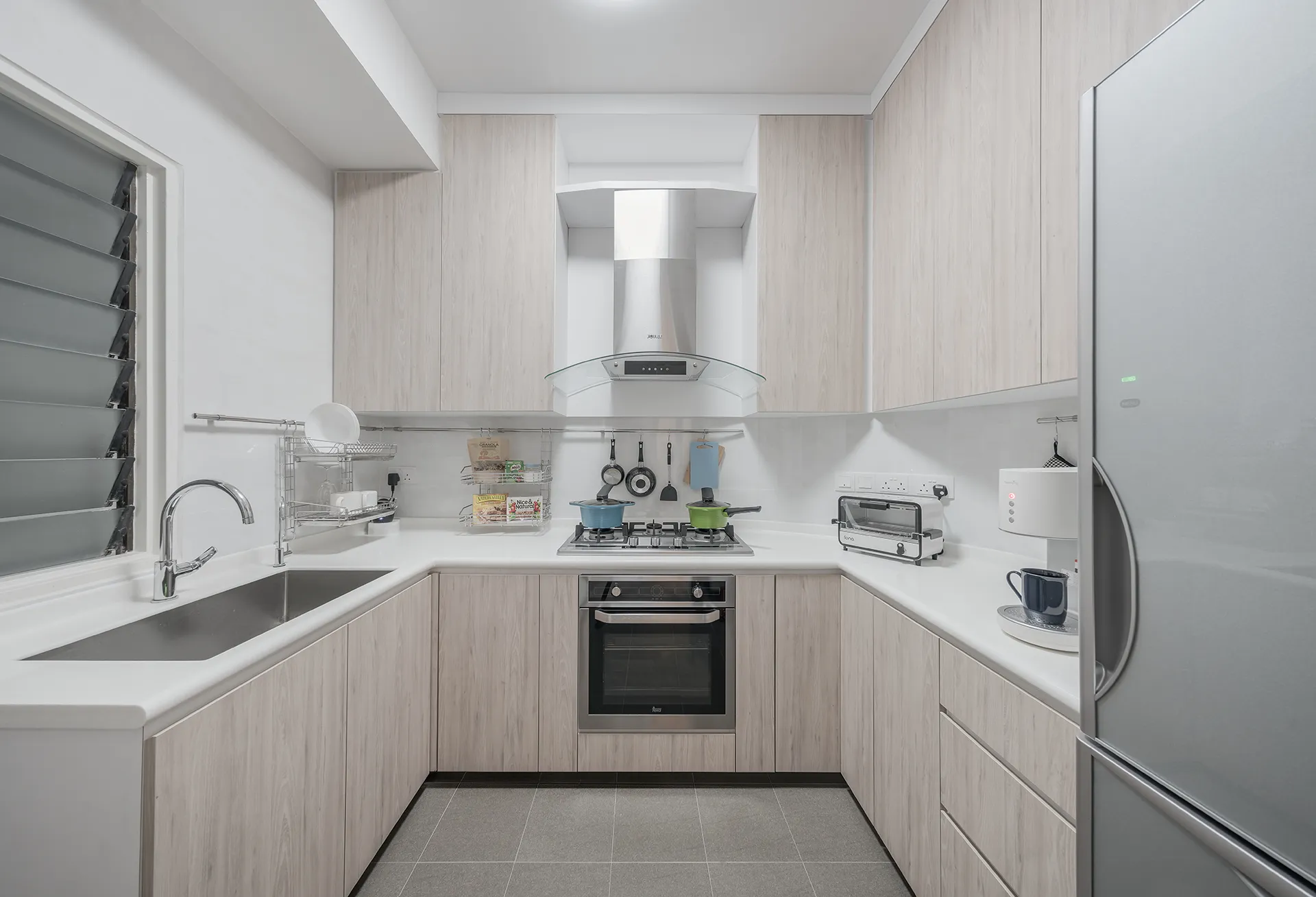 A modern galley kitchen with a linear layout, featuring light wood base cabinets and a white countertop. A stainless steel sink and a two-burner gas stove are positioned under a window, showcasing a clean and efficient design.