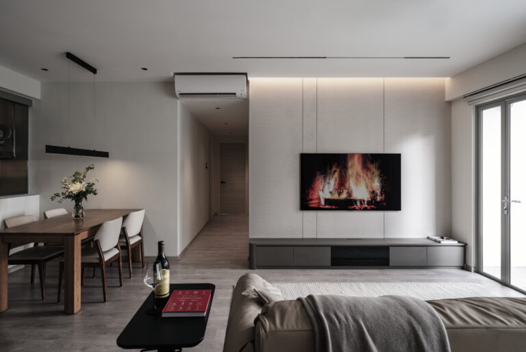 Modern contemporary living room with a wall-mounted TV displaying a fireplace, grey entertainment unit, beige sofa, black side table, and wooden dining table with white chairs.