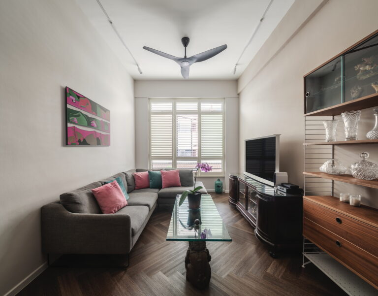 Modern living room with grey sectional sofa, colourful pillows, glass coffee table, TV, ceiling fan, and decorative shelves.