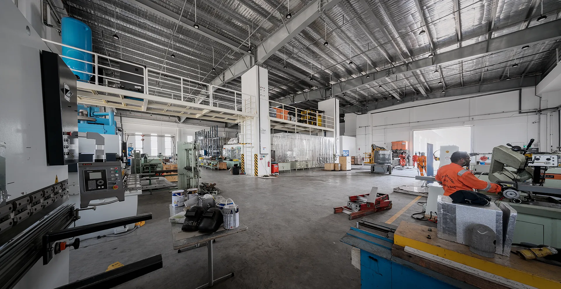A large, high-ceilinged industrial workshop with heavy machinery, workbenches, a mezzanine level, and a worker in a high-visibility jacket.
