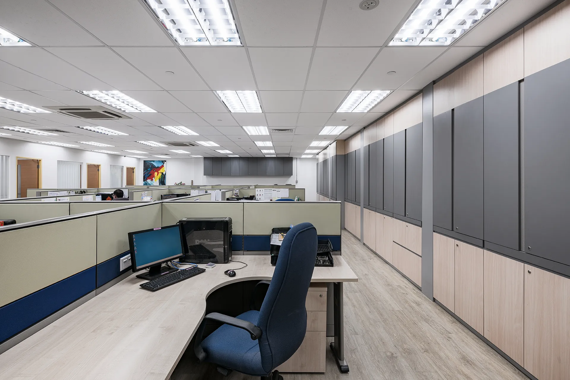 An open-plan office space with rows of modular workstations, built-in storage units, and a colorful abstract painting on the back wall.