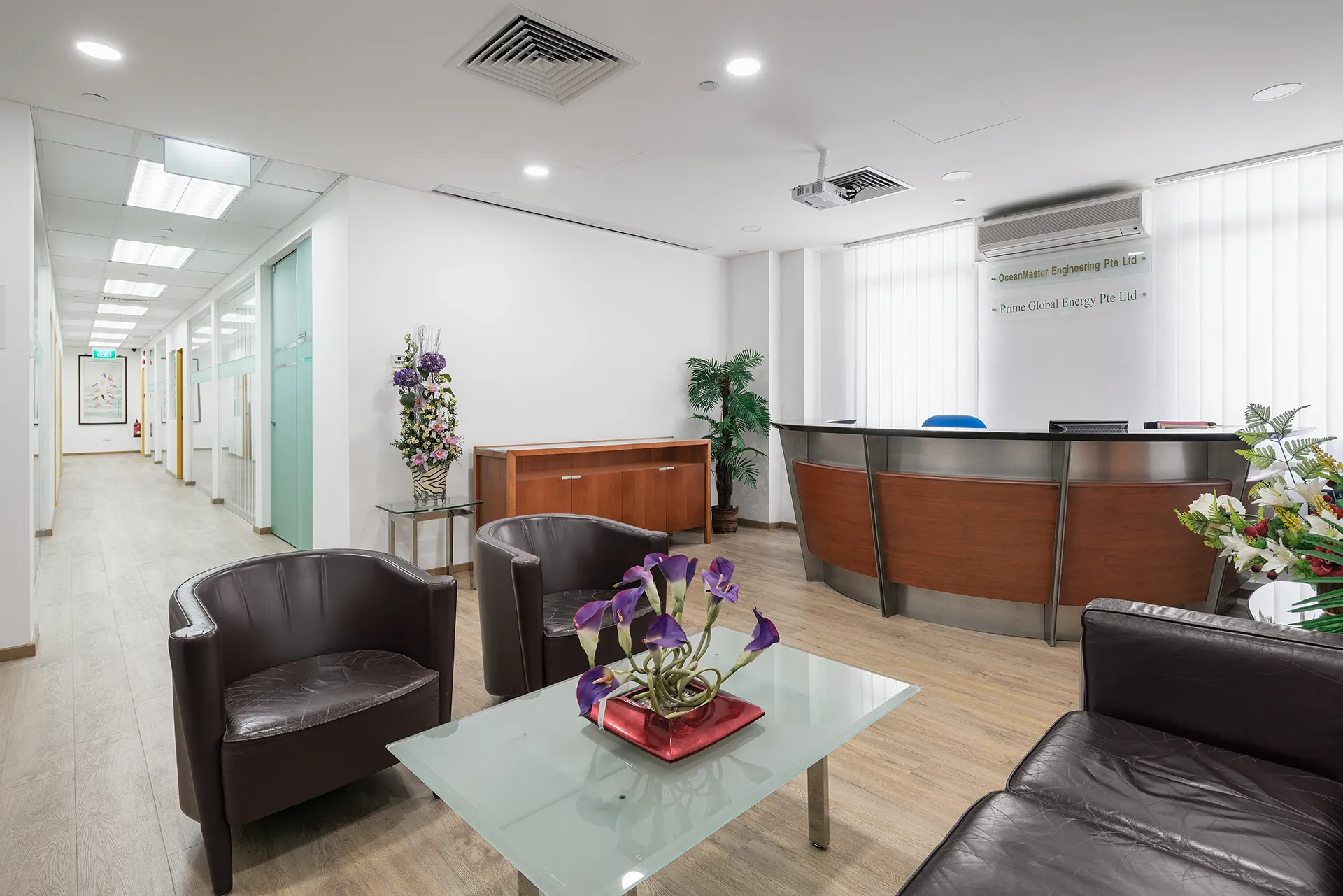 A modern, professional office reception area with a central desk, two brown leather armchairs, a glass coffee table, and signage for "OceanMaster Engineering Pte Ltd" and "Prime Global Energy Pte Ltd."