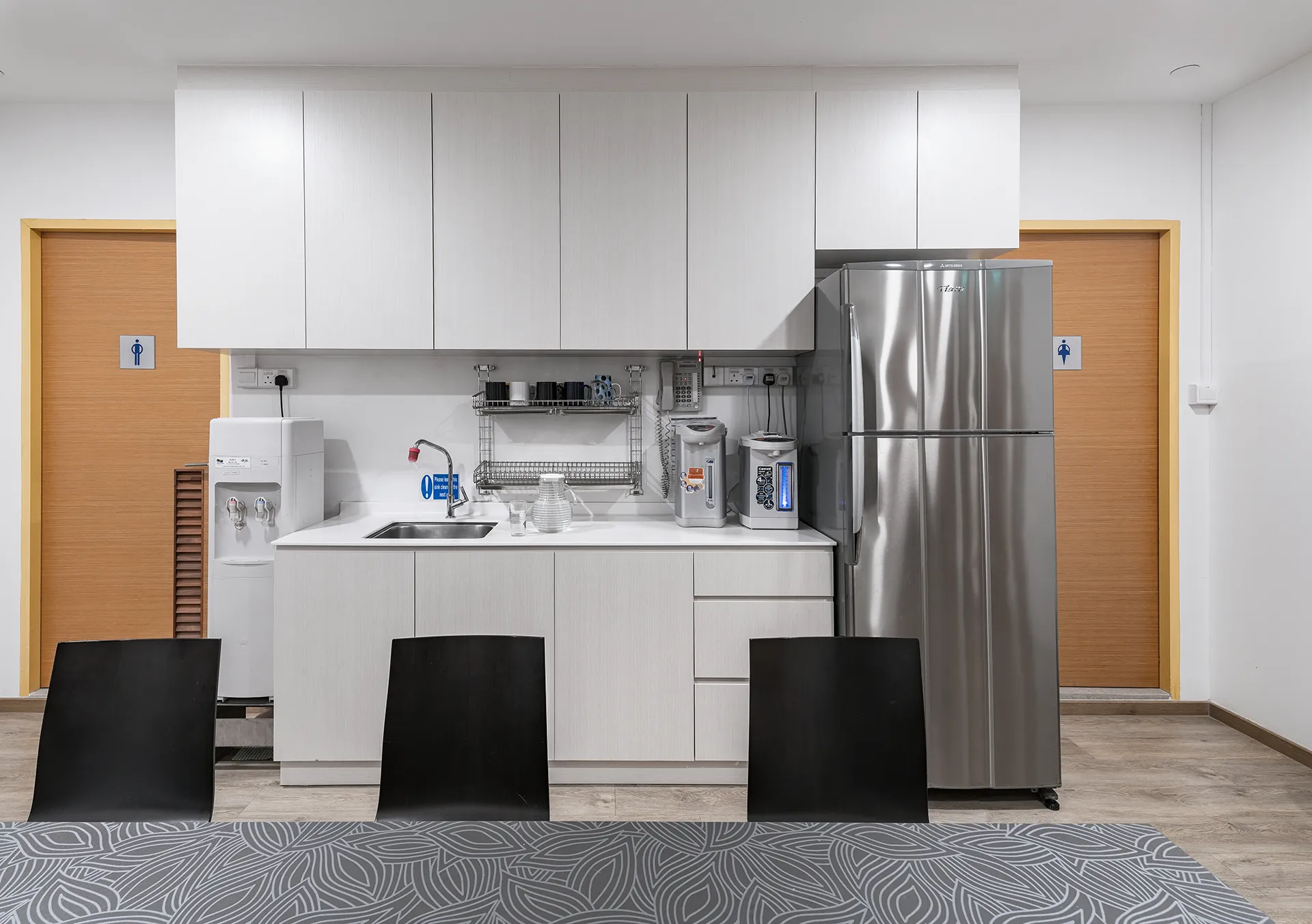 A clean, modern office canteen with a patterned tablecloth on a table, black chairs, white cabinetry, a stainless steel refrigerator, and a water dispenser.