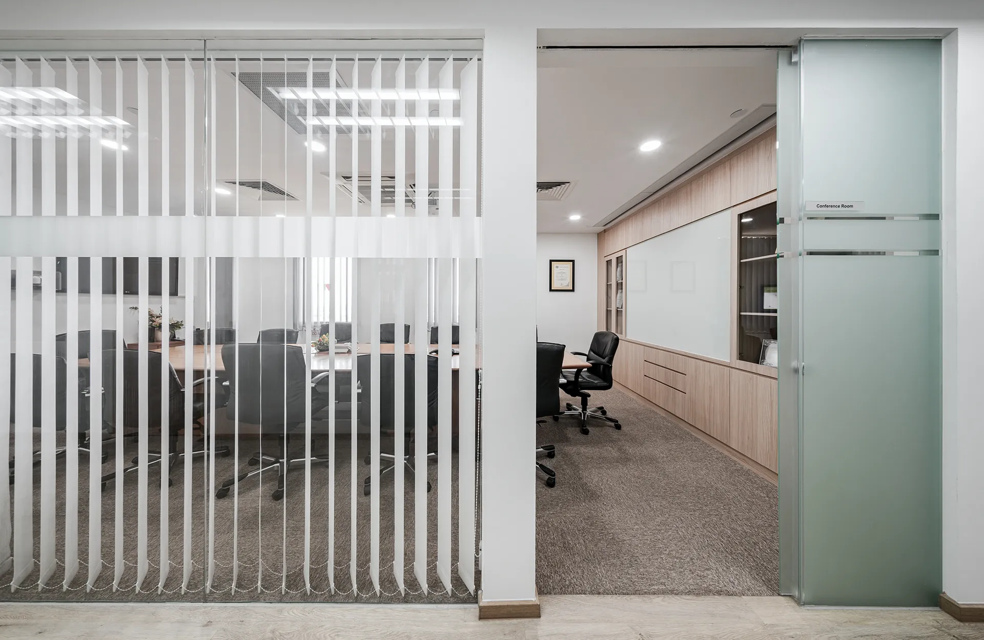 A view of two adjacent conference rooms separated by a frosted glass partition, featuring black ergonomic chairs, large tables, and built-in wooden cabinetry.
