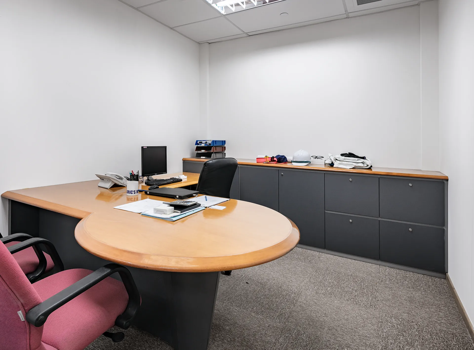 A compact private office with a U-shaped wooden desk, grey cabinetry, a black and a red office chair, and fluorescent lighting.