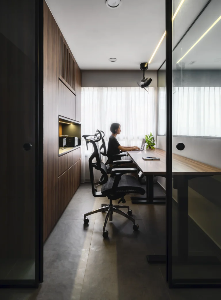 Scandinavian office space with a person working at a desk, ergonomic chairs, wooden cabinetry, and large windows with sheer curtains.