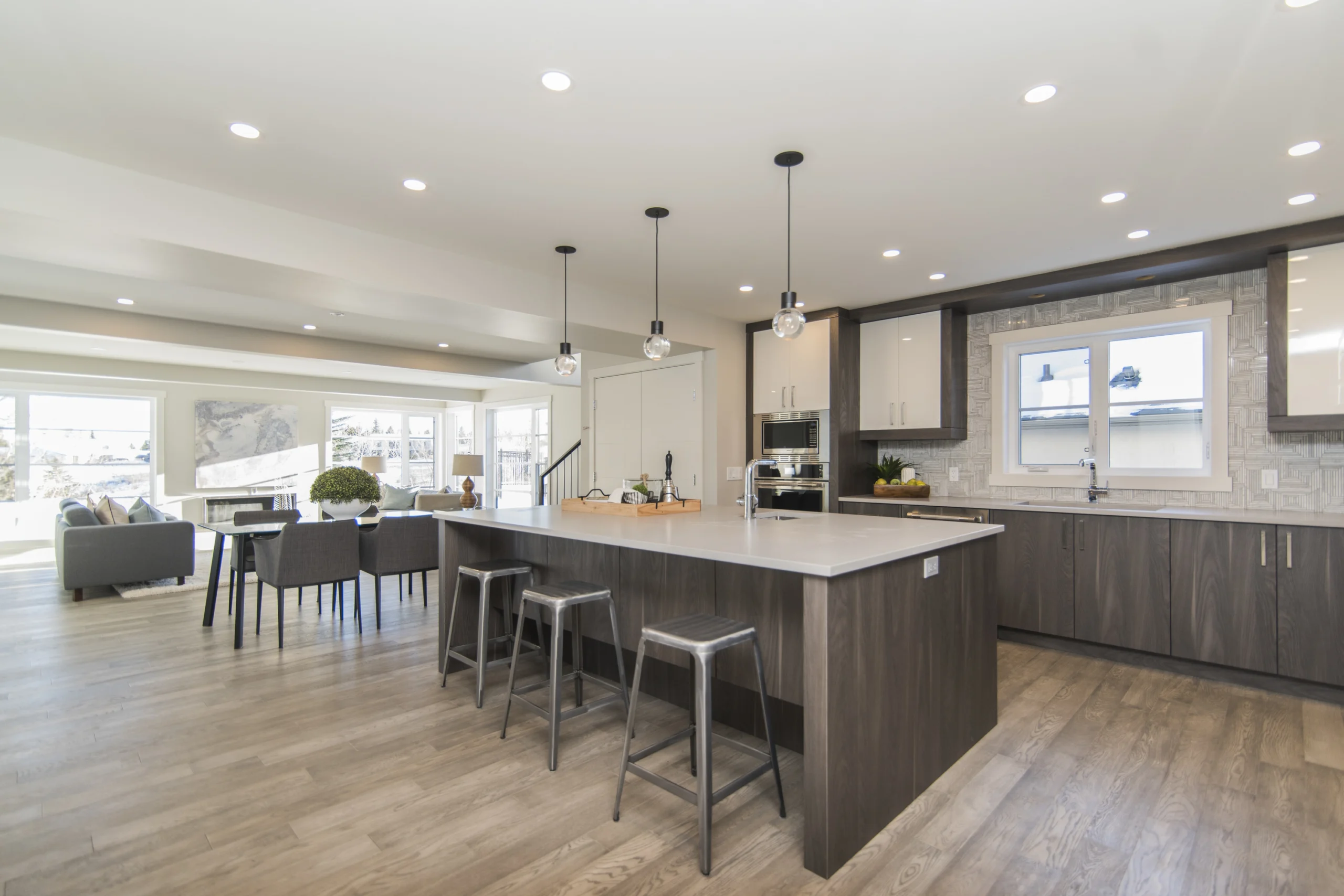 Modern open-plan kitchen and living area with large island, metal bar stools, pendant lights on HDB flat's ceiling, dark wood and white cabinets, built-in appliances, and dining table.