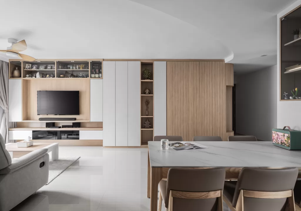 A Contemporary Scandinavian living room and dining area with a minimalist design. The living room features a wall-mounted TV with a wooden and white storage unit that includes shelves with various decorative items and books. There is a light-colored sofa and a white coffee table with a tissue box on it. The dining area has a white marble table with four beige chairs, a mug, and an open magazine on the table. A ceiling fan with wooden blades is visible on the left side of the image. The floor is tiled in a light color, and the overall decor is neutral with wooden accents.