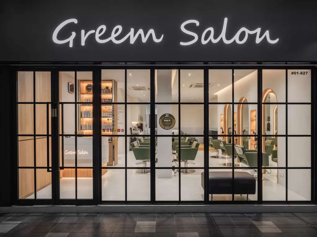 Exterior view of Greem Salon with large glass windows and doors, showcasing the interior with green salon chairs, mirrors, and a reception desk. The salon name 'Greem Salon' is prominently displayed in illuminated letters above the entrance.