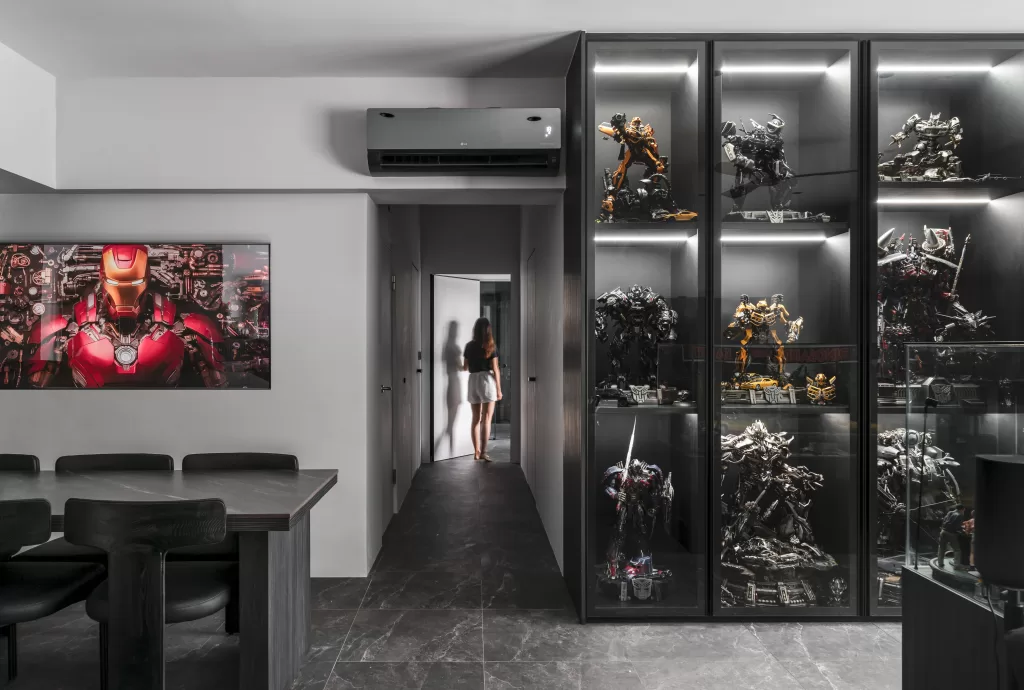 A modern room featuring glass cabinets filled with a collection of intricate robot figurines on the right side, reflecting a passion for sci-fi or superheroes. On the left, a sleek dining table with black chairs sits beneath a large, striking Iron Man artwork that dominates the wall. An air conditioning unit is mounted above the doorway, and a person walks through the doorway in the background, adding a sense of scale and casual ambiance to the space.