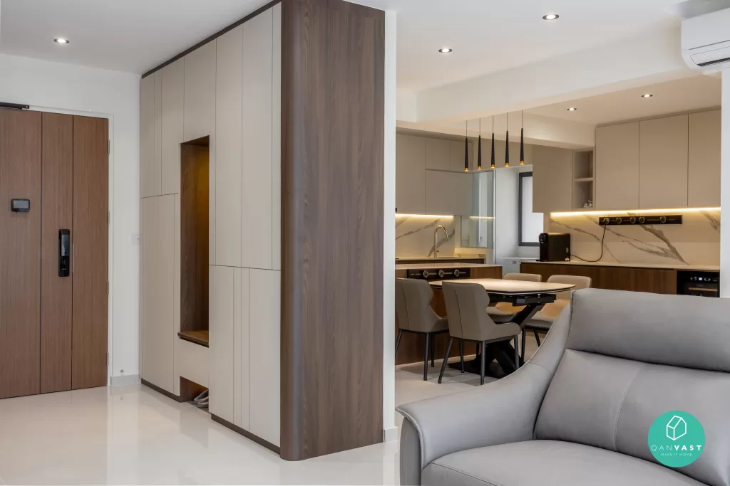 A modern contemporary, minimalist interior of a home featuring a kitchen and dining area. The kitchen has sleek cabinetry with a marble backsplash and under-cabinet lighting. The dining area includes a table with four upholstered chairs. A gray leather sofa is partially visible in the foreground. The entrance door and built-in storage cabinets are on the left side of the image. The room is well-lit with recessed ceiling lights.
