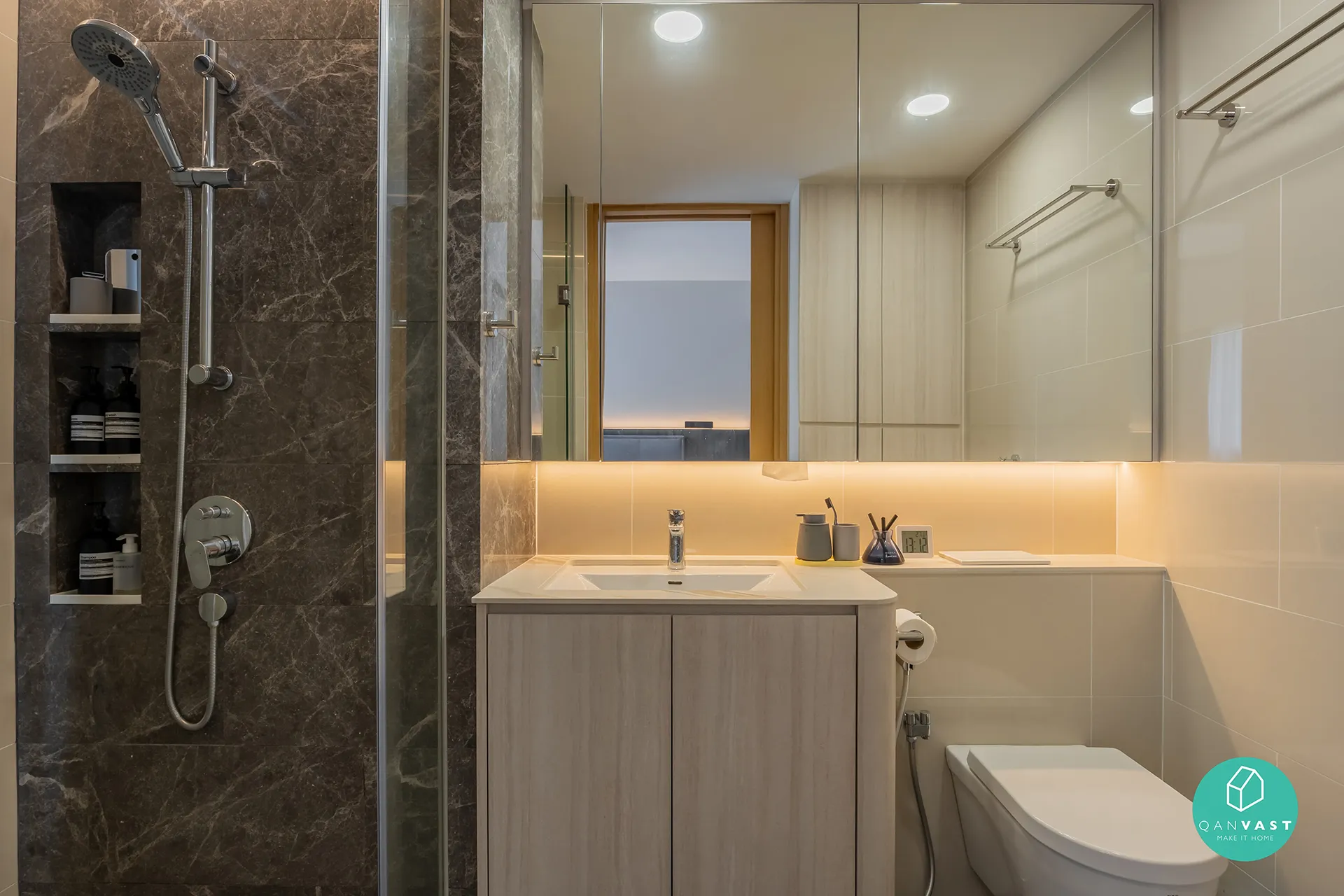A luxurious bathroom interior at Bartley Ridge, Singapore, showcasing a dark marble accent wall with a built-in shelf in the shower. The image highlights a glass partition, light wood vanity, and a large mirror with warm, integrated lighting.