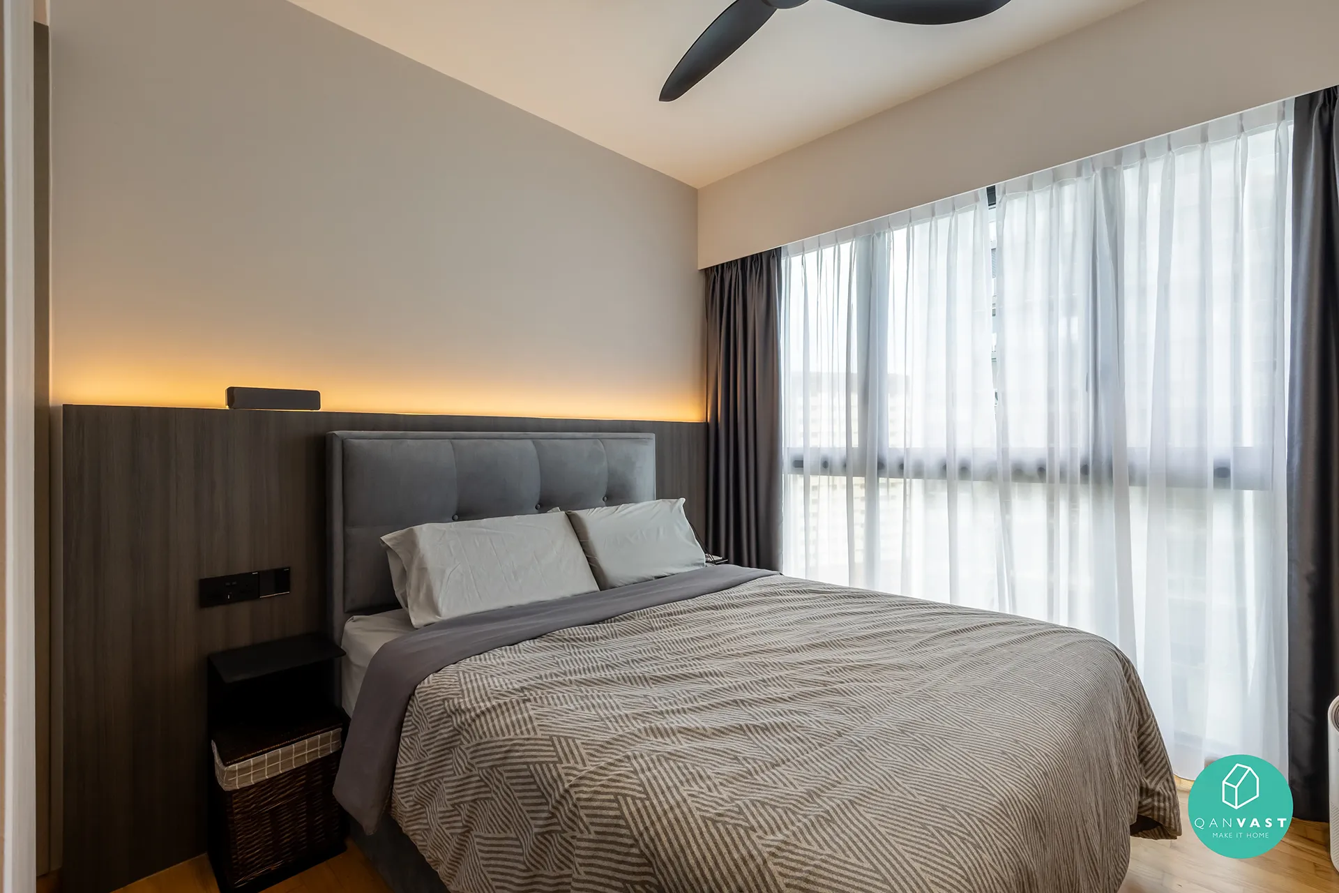 A tranquil and minimalist bedroom at Bartley Ridge with a grey upholstered headboard and layered bedding. The image highlights a recessed strip of warm ambient lighting, a compact nightstand with a lamp, and sheer curtains over a large window.
