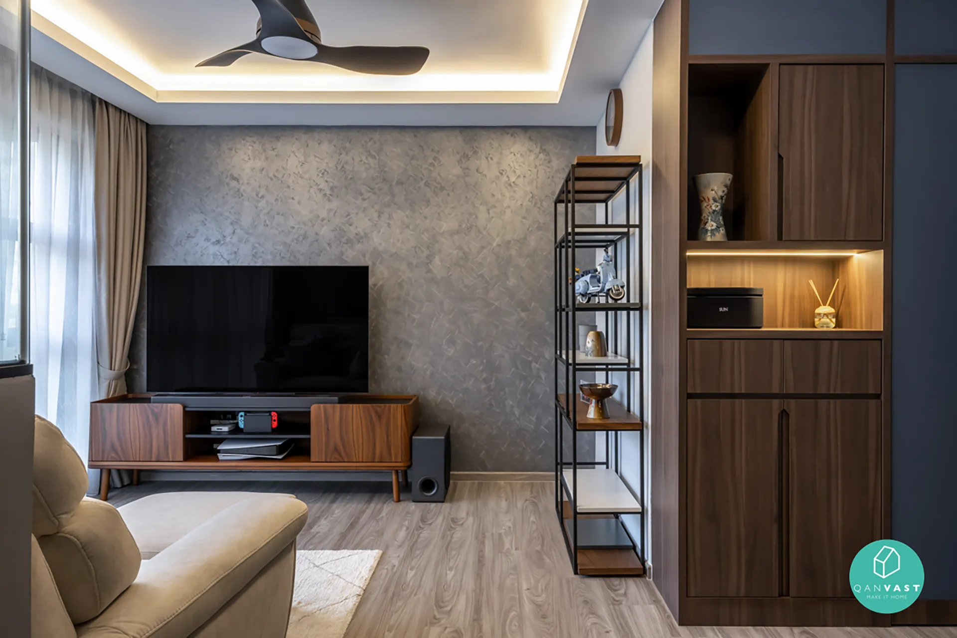 Modern living room with TV entertainment wall, textured grey backdrop, beige sofa, shelving with decorative items, and light wood flooring.