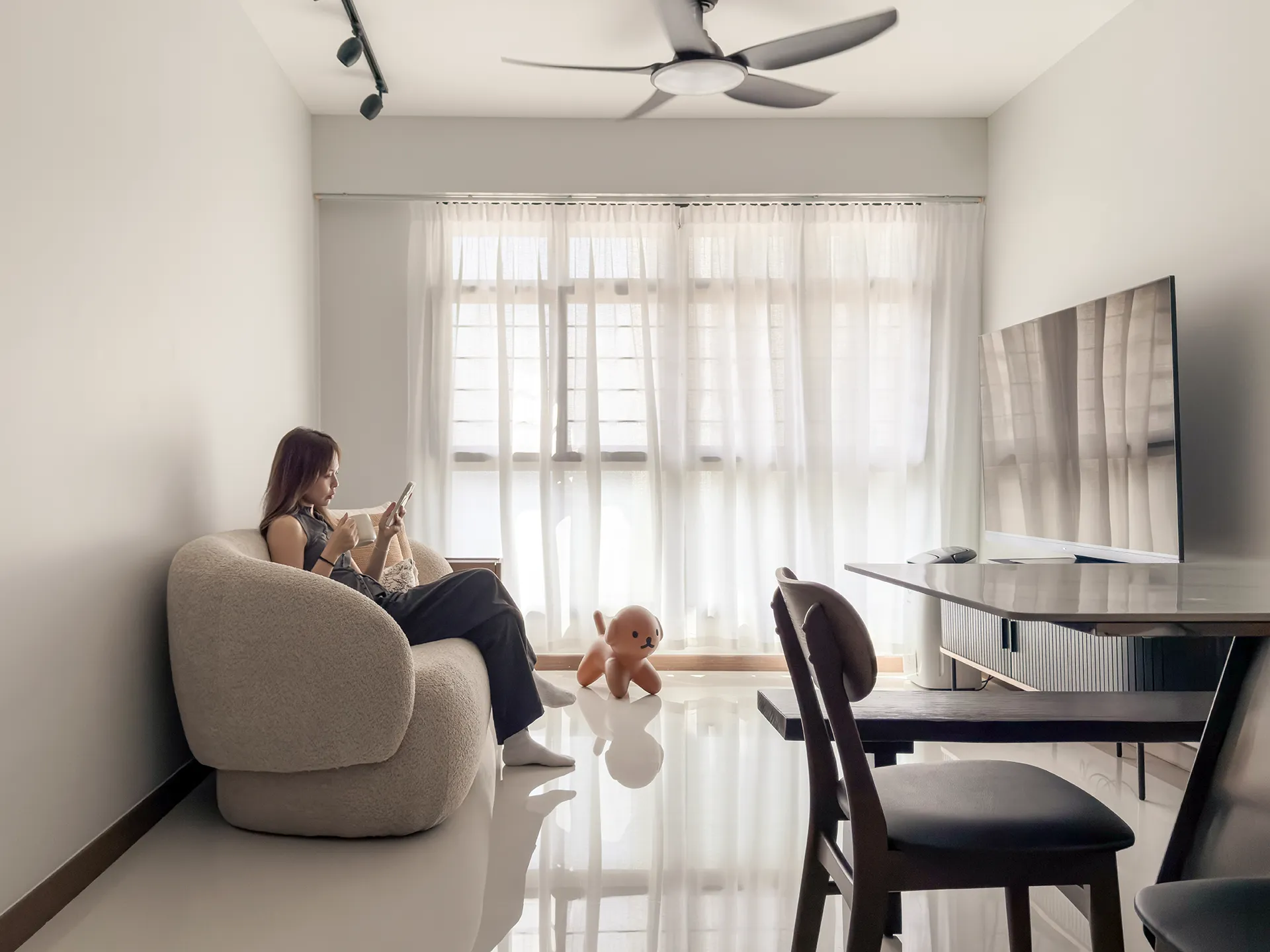 Bright living room with large floor-to-ceiling window, sheer white curtains, beige armchair with occupant holding tablet, oversized brown dog toy on polished floor, dark wooden desk and chair, and ceiling fan.