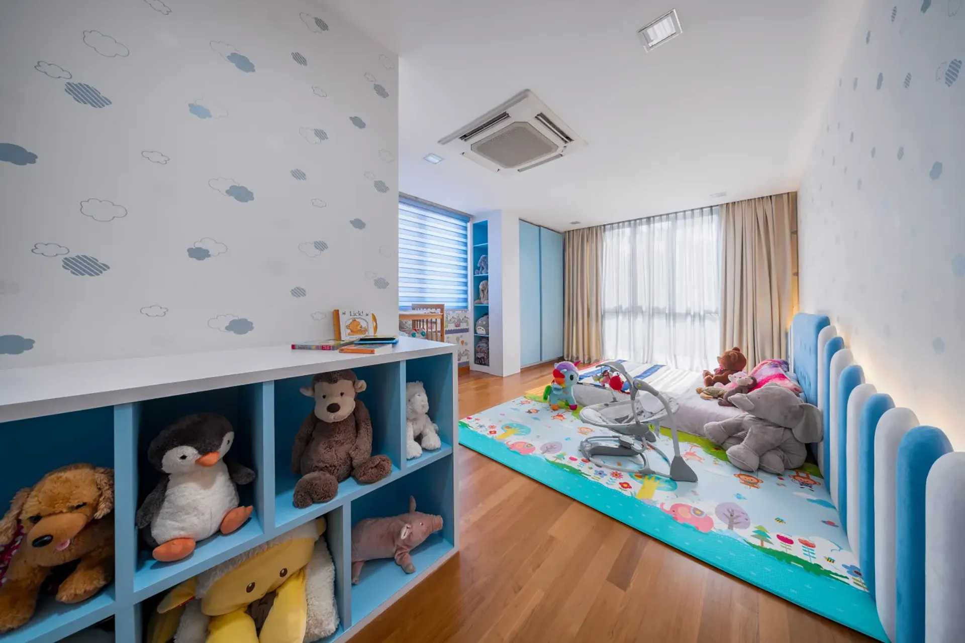 A bright, playful nursery and playroom in a Singapore home with cloud-patterned walls, a white crib, a padded blue headboard, and a vibrant animal-themed play mat on a wooden floor.