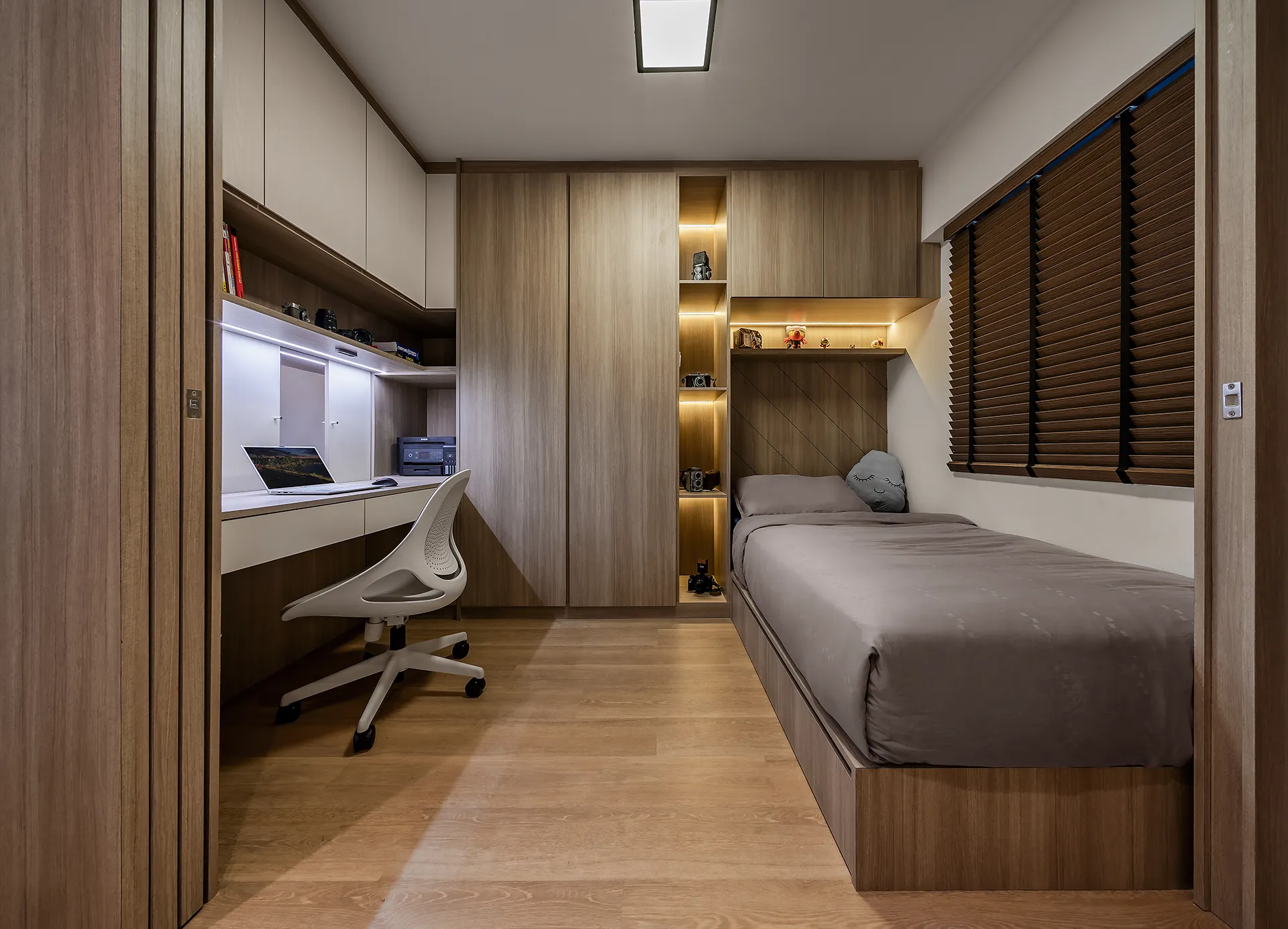 Minimalist HDB bedroom and study area with built-in light wood desk, floating shelves displaying vintage cameras, and gray duvet single bed at 427 Clementi Avenue 3.