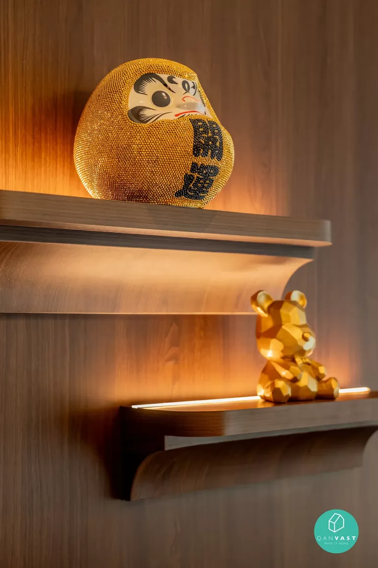 Detail of cultural accents on a warm-lit wooden shelf in a One Pearl Bank condo, Singapore, showcasing a golden Daruma doll and a faceted bear figurine.