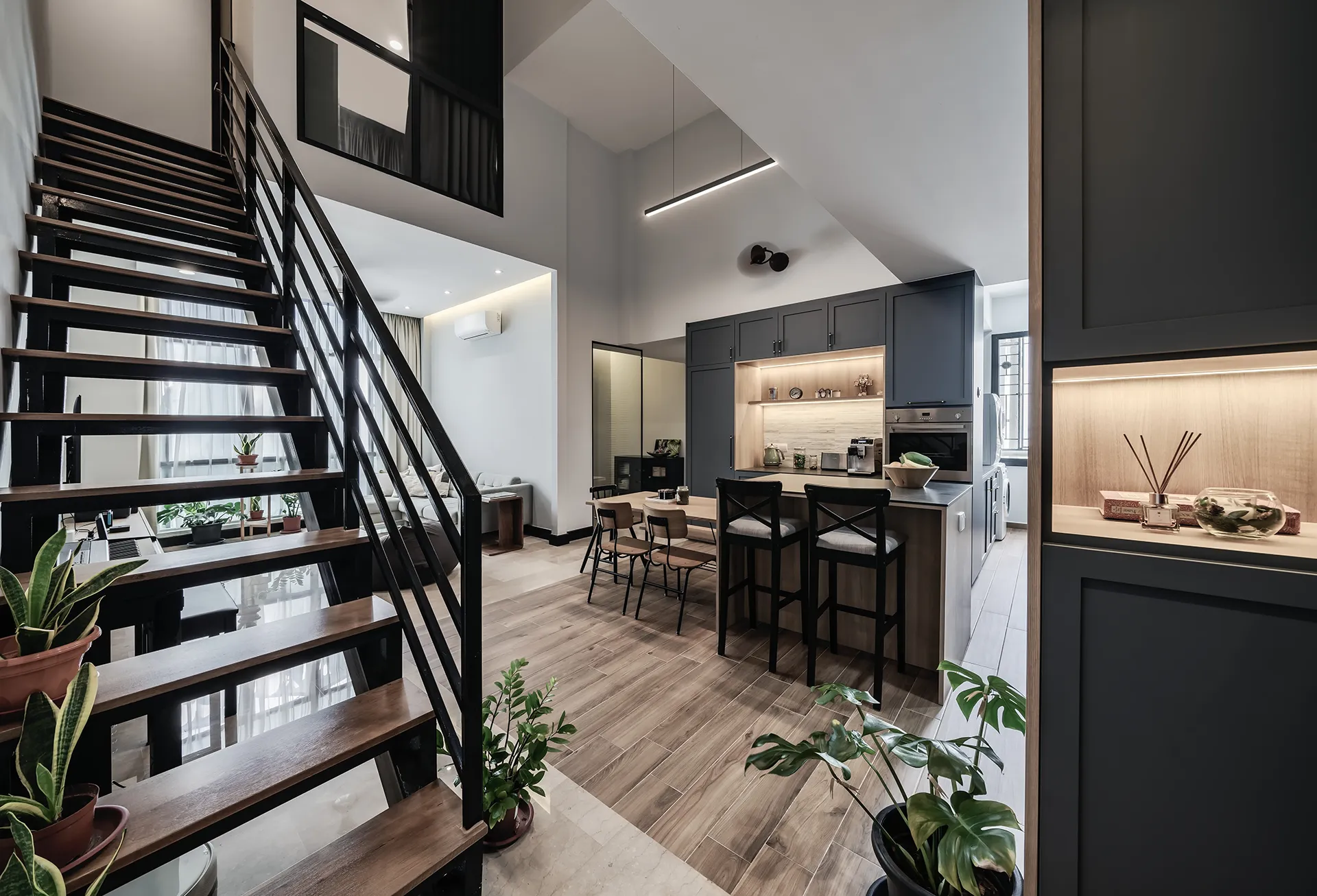 A contemporary, open-concept kitchen and living area in Singapore with dark matte cabinetry, a light wood floor, and a sculptural black and wood staircase. The central island has bar seating for three.