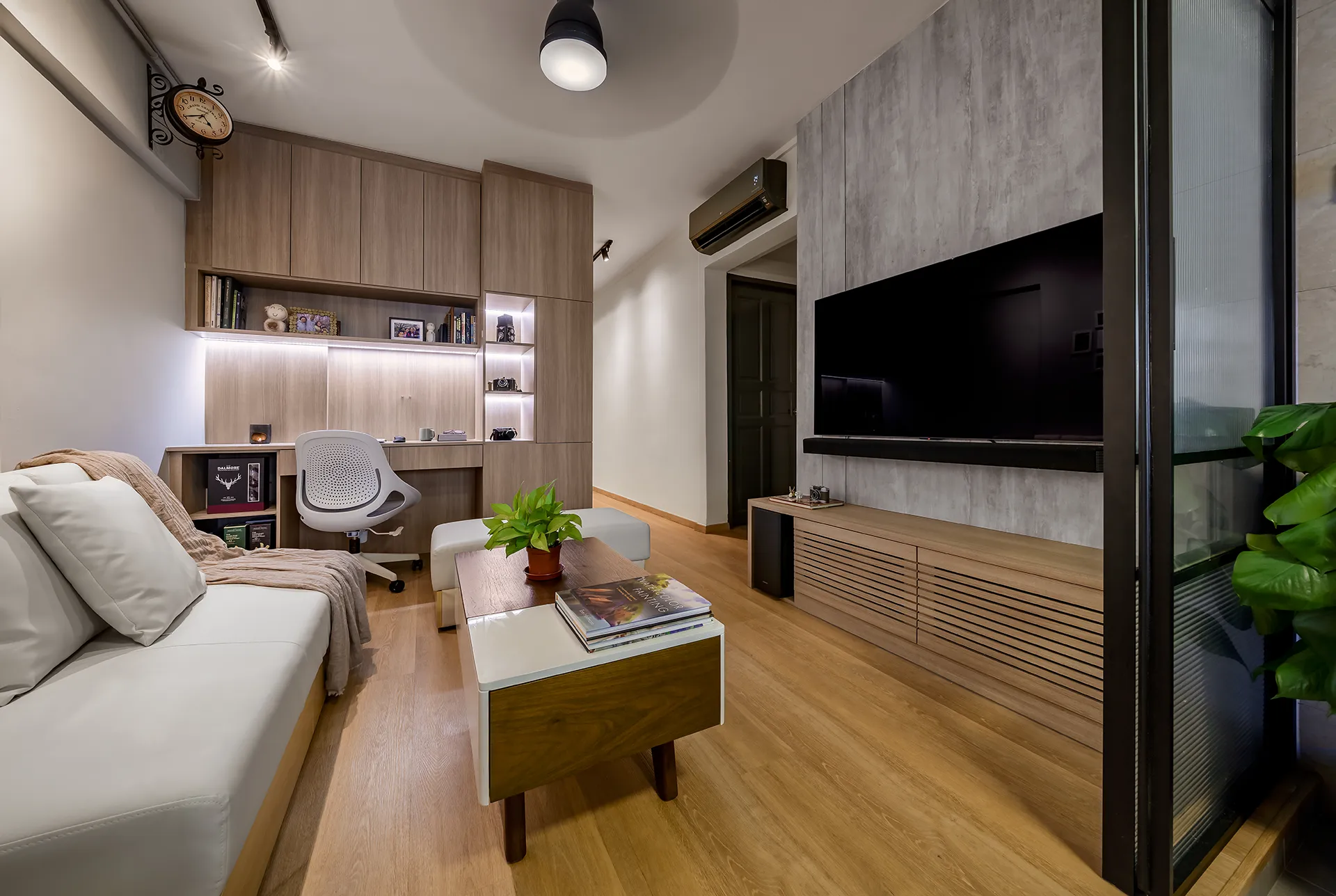 Open living and lounge area with white sofa, built-in wood storage and work desk, concrete TV wall, and glass door leading to balcony in Clementi HDB flat.