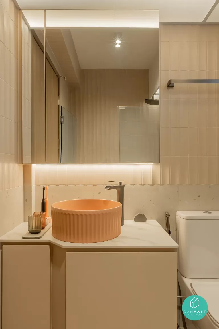Modern HDB common bathroom with peach ribbed vessel sink, vertical beige tiles, and large mirror with integrated warm ambient lighting.