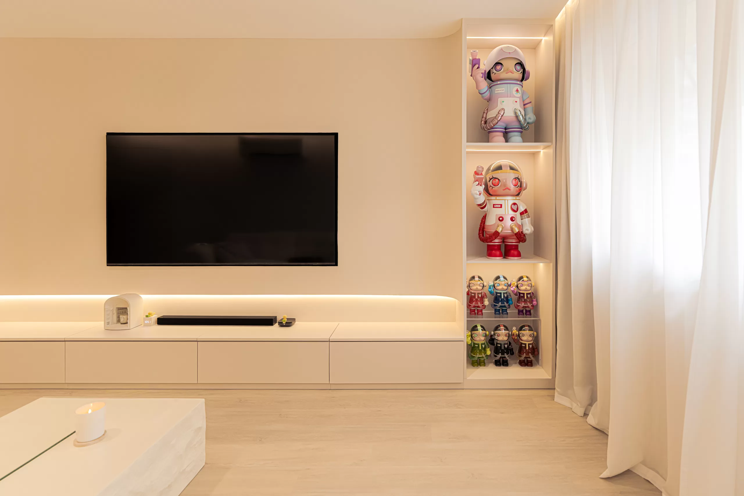 Minimalist 4-room HDB living room with a beige accent wall, low-profile media console, and a recessed, illuminated shelf displaying colorful astronaut figurines.