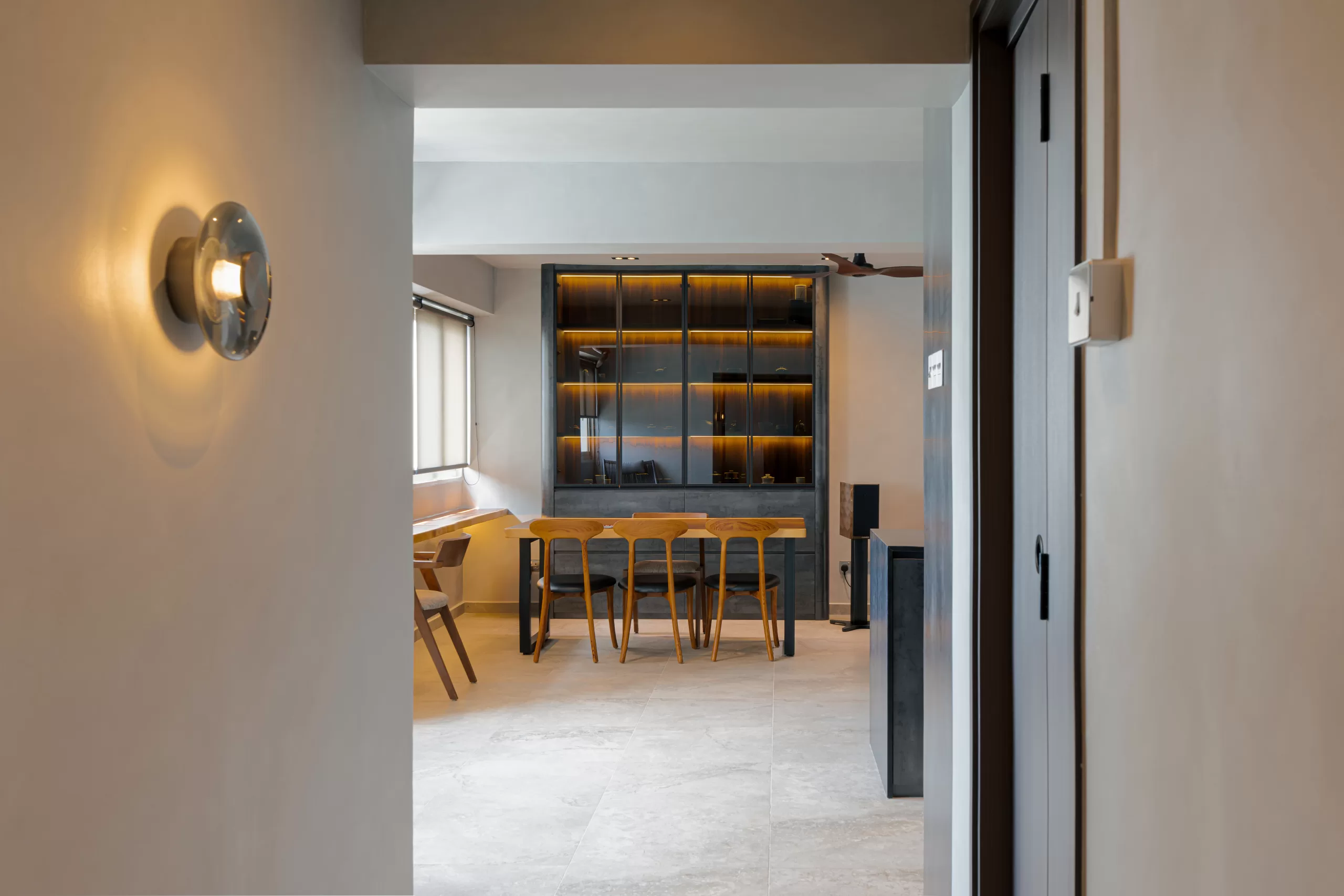 Transitional view into a multifunctional HDB room blending dining and study functions, anchored by a wooden table and a backlit glass-fronted display cabinet.