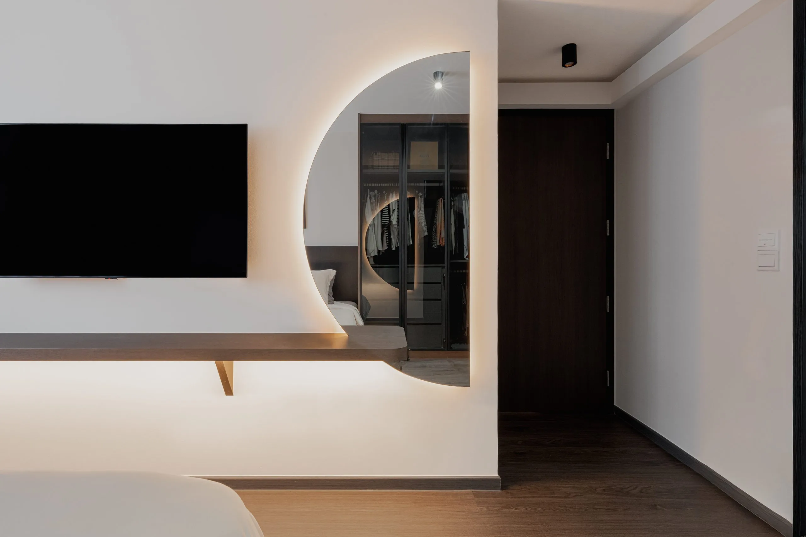 Bedroom wall with a wall-mounted TV, a floating shelf and a backlit half-round mirror