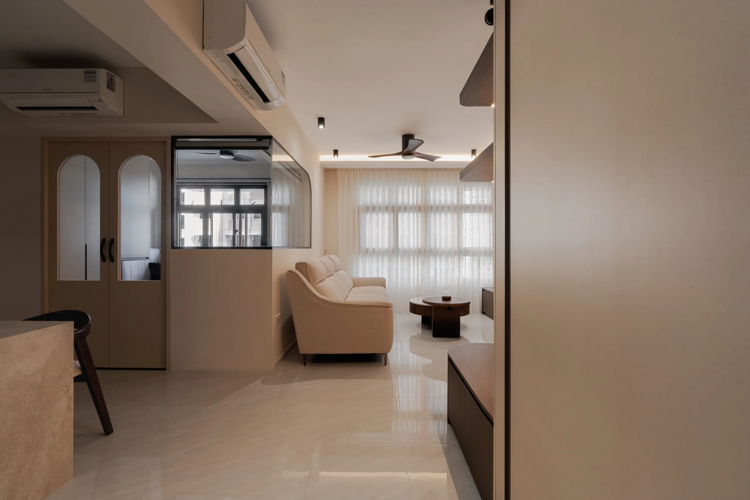 Living room with a cream sofa and glossy floor tiles, next to a half-height wall topped with a glass panel and arched double doors