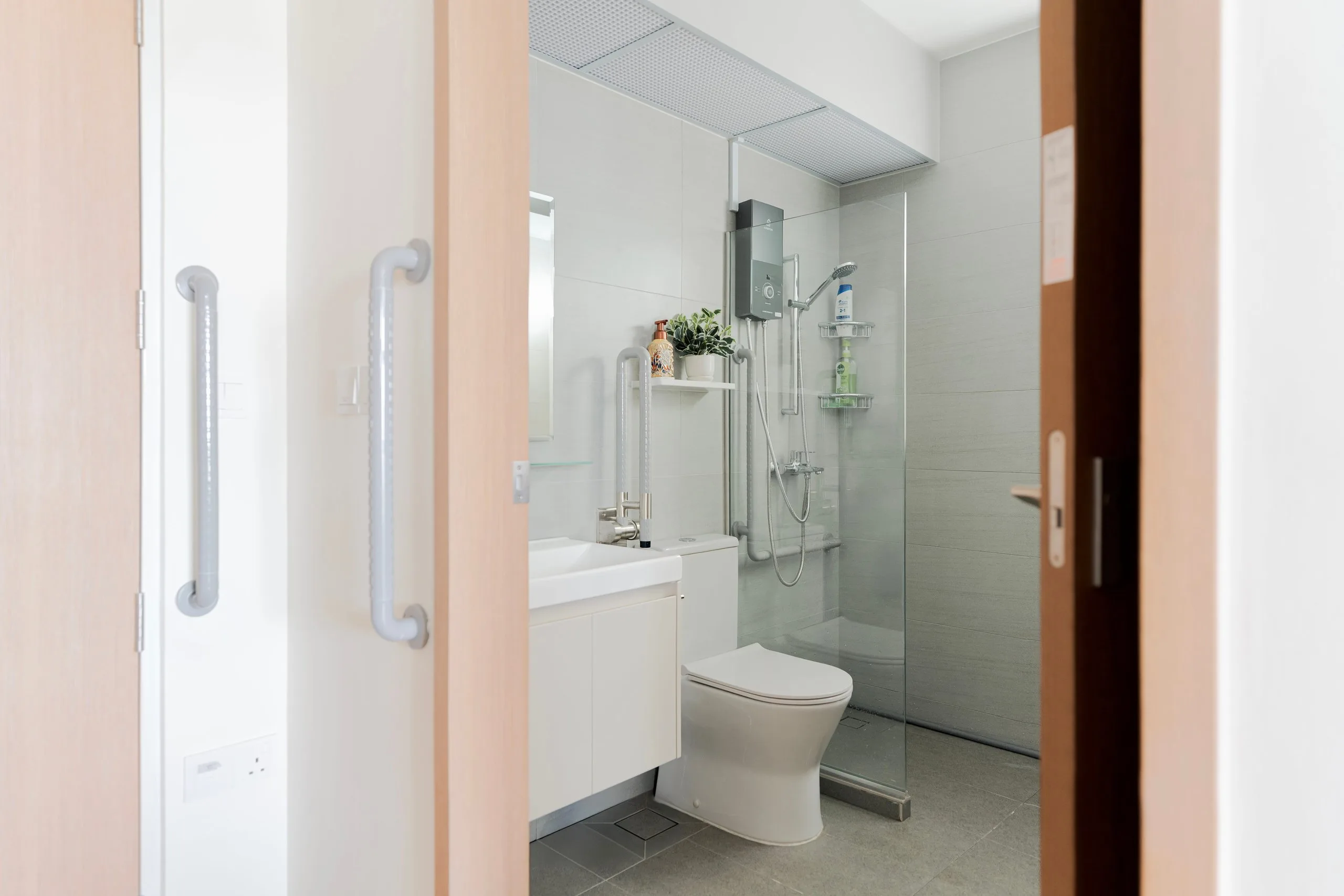 Bathroom with a white vanity, a glass shower screen, wall shelves and grab bars, seen through a sliding door