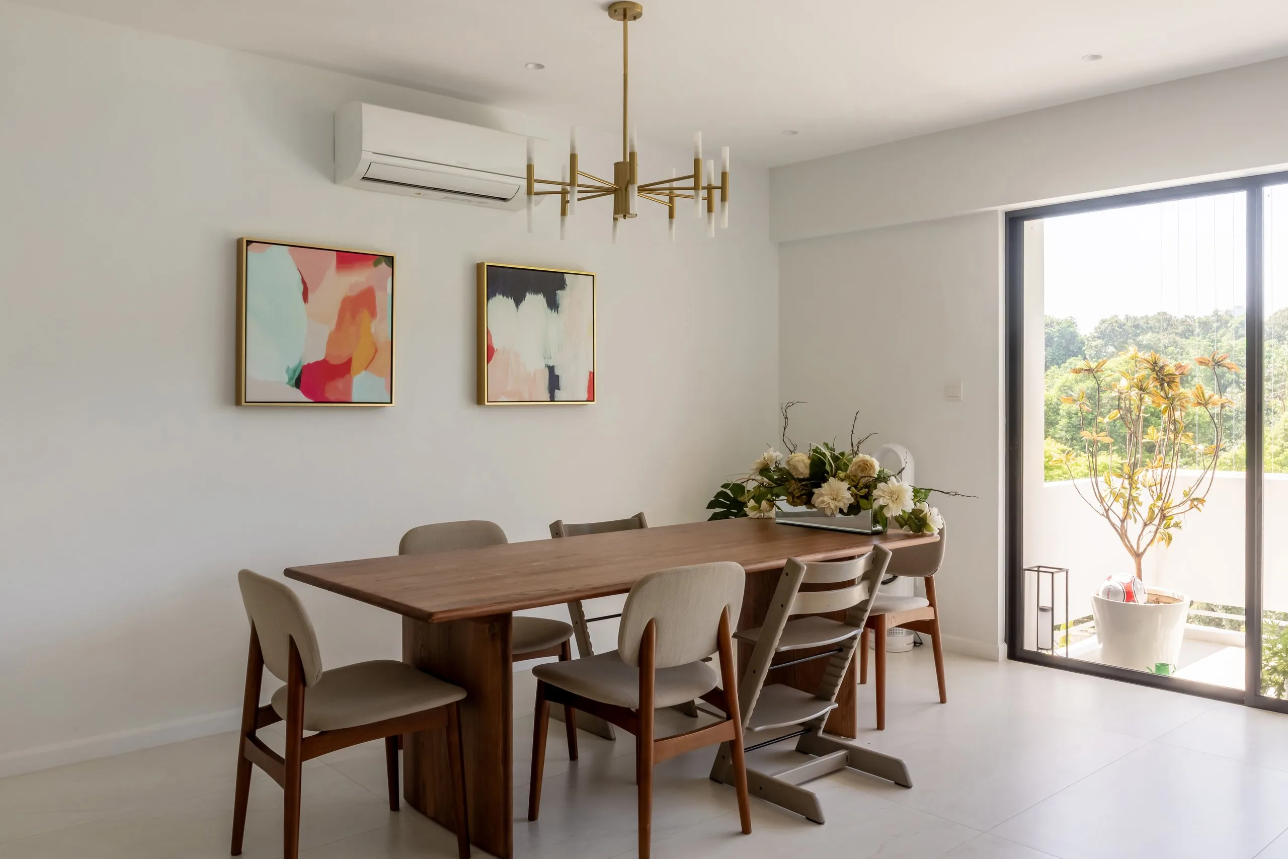 Wood-toned dining table and chairs under a brass-toned chandelier, beside full-height glazing with greenery outside