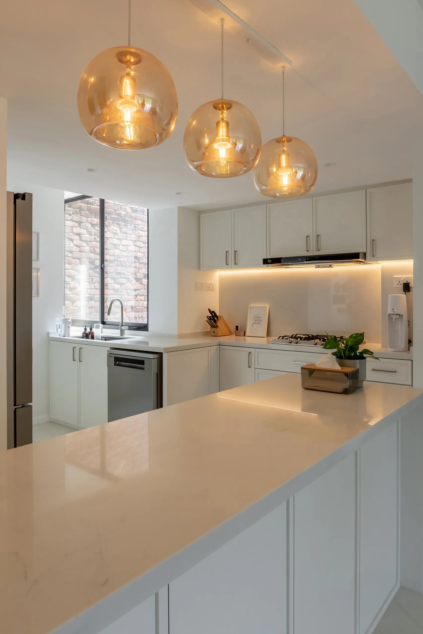 White kitchen peninsula with a lightly veined worktop under three amber glass pendants, with framed white cabinets and under-cabinet lighting behind