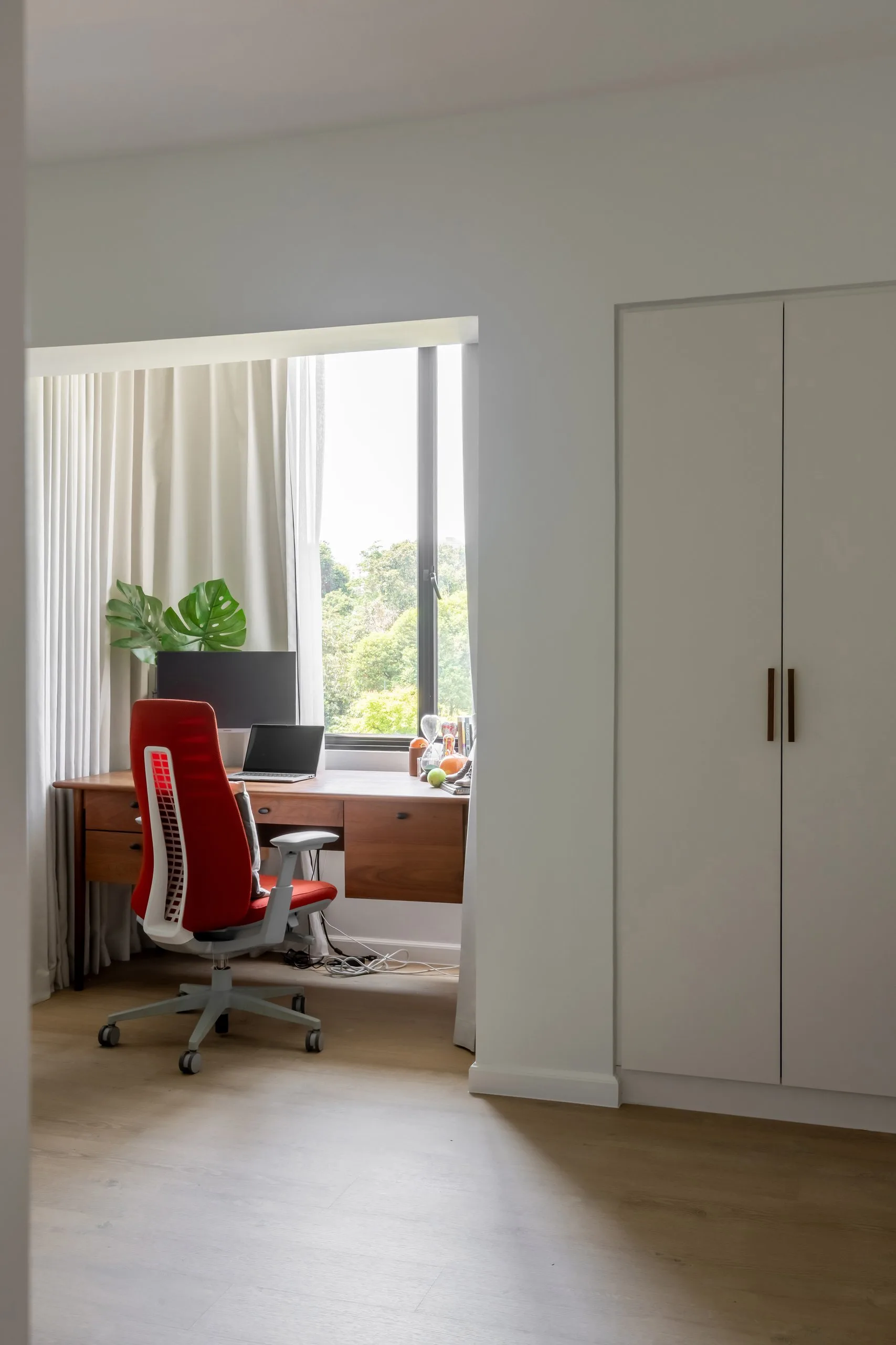 Wood-toned desk and red office chair set into a window recess with a view of trees, next to a white built-in wardrobe